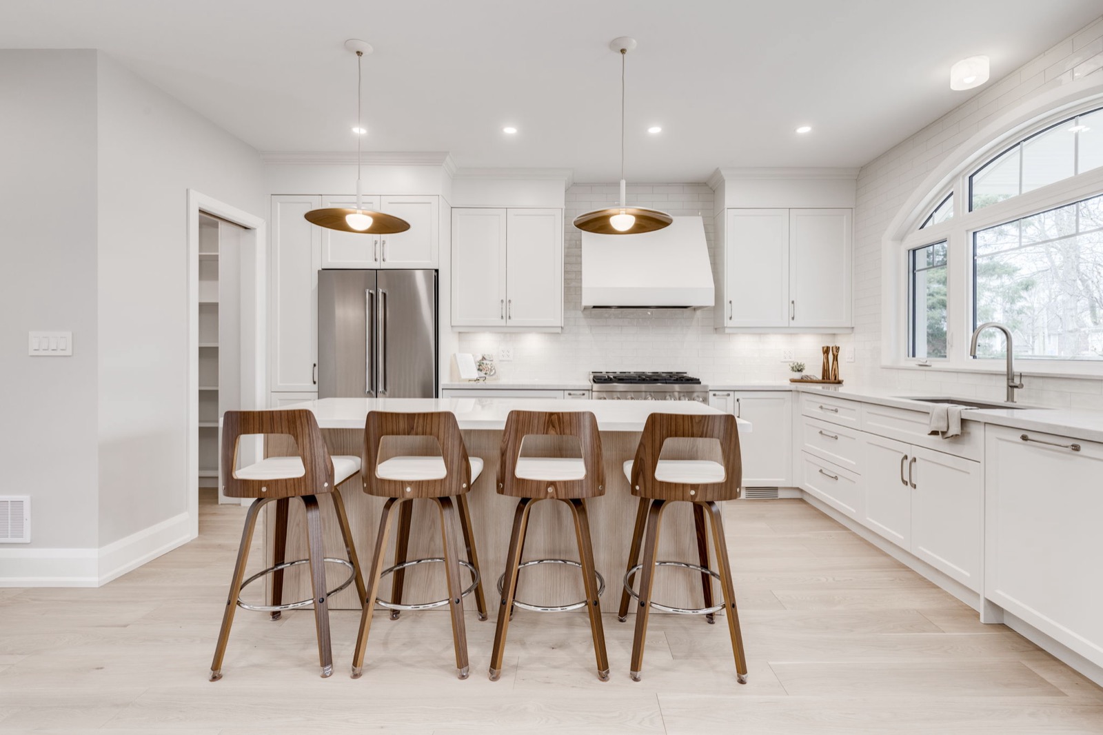 Bright white kitchen renovation with a large island, wood stools, arched window, and clean tile backsplash.